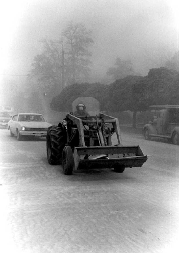 Black and white image of bulldozer in the middle of the street being follow up a line of cars. Driver of bulldozer is wearing protective gear to protect against the ash. 