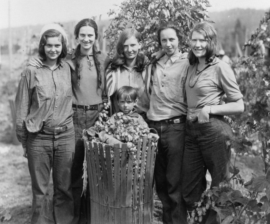 Five young women are standing behind a basket of hops. In the basket is a little boy with hops on his lap (Source: Trover Studio).