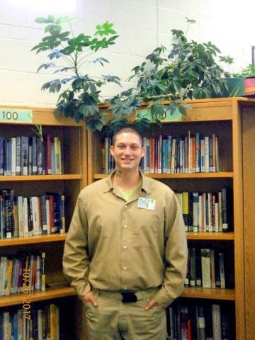 A young man in a prison uniform standing in front of a shelf of books. Healthy houseplant on top of the bookcase.